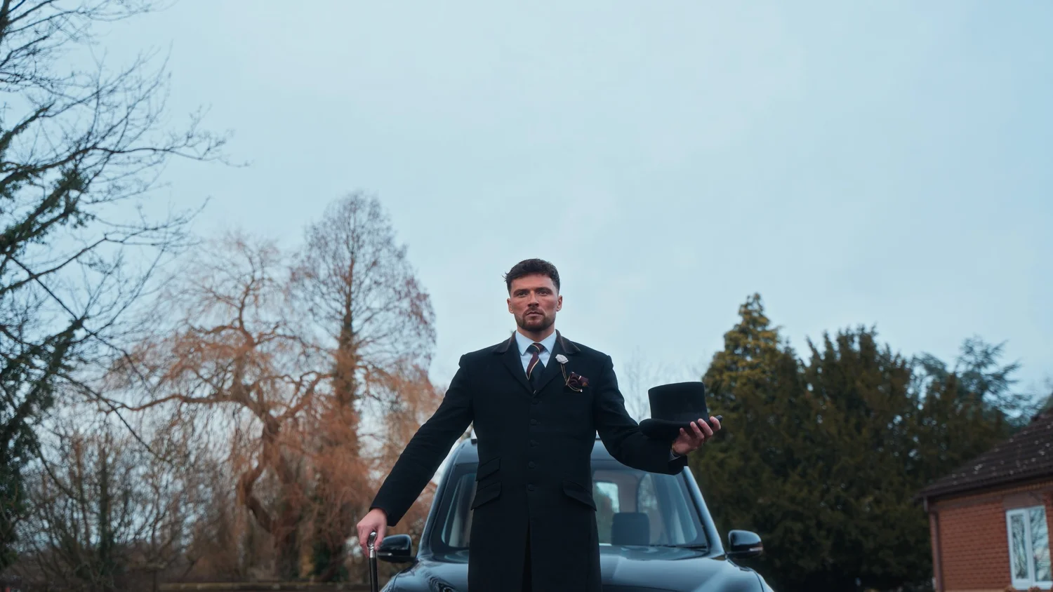 A man in vintage formalwear standing beside a classic black car.